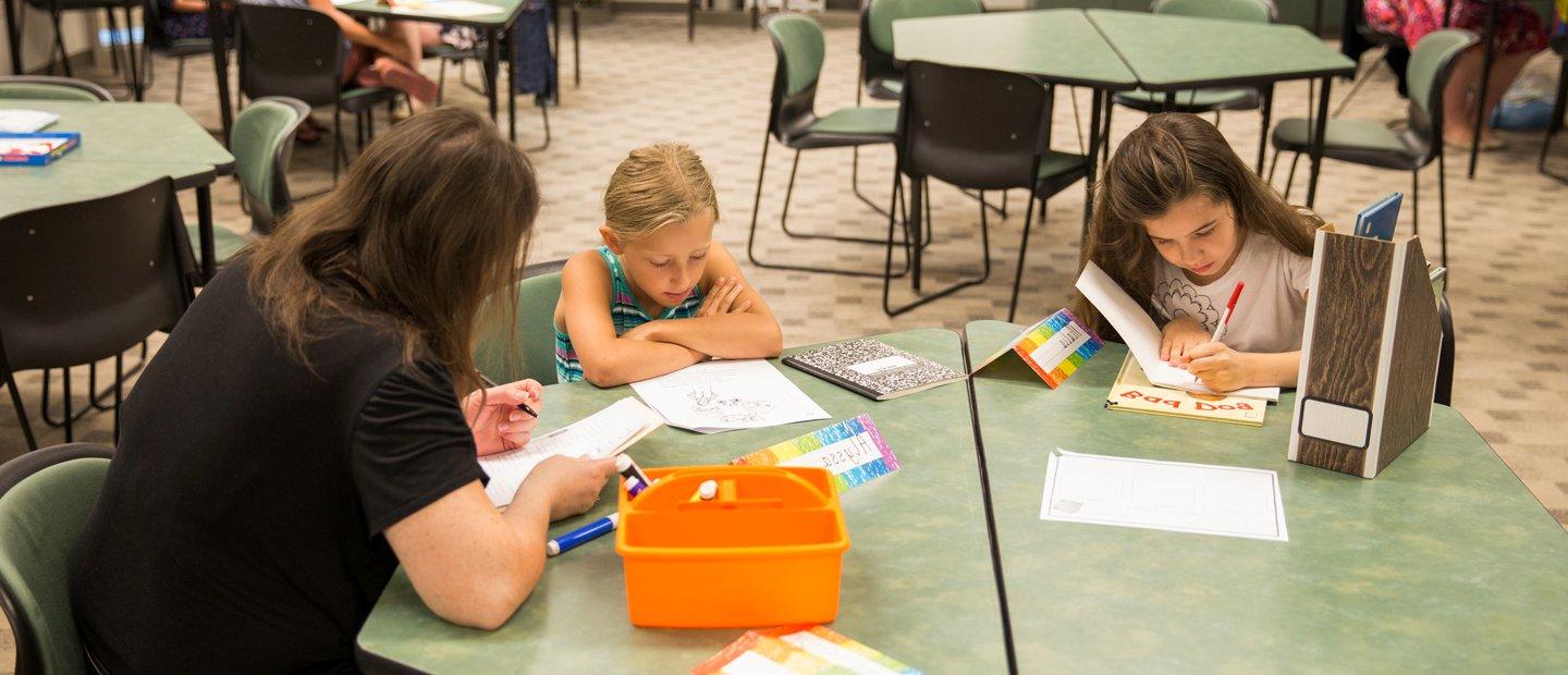 Two young girls sitting with a woman at a green table, writing and reading.